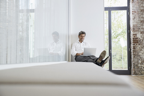 Businessman sitting on bench in modern office using laptop stock photo