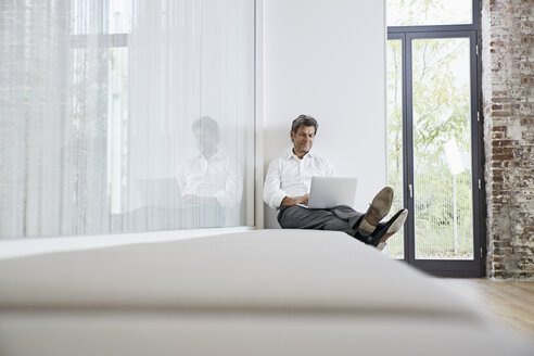 Businessman sitting on bench in modern office using laptop - PDF01458