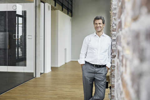 Porträt eines lächelnden Geschäftsmannes, der sich in einem modernen Büro an eine Backsteinwand lehnt, lizenzfreies Stockfoto