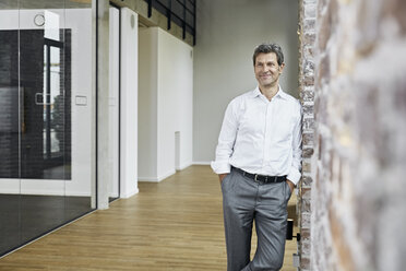 Porträt eines lächelnden Geschäftsmannes, der sich in einem modernen Büro an eine Backsteinwand lehnt - PDF01469