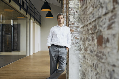 Portrait of confident businessman leaning against brick wall in modern office - PDF01474