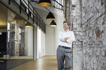 Portrait of smiling businessman leaning against brick wall in modern office - PDF01475