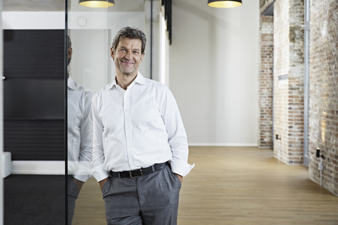 Portrait of smiling businessman leaning against glass pane in modern office stock photo