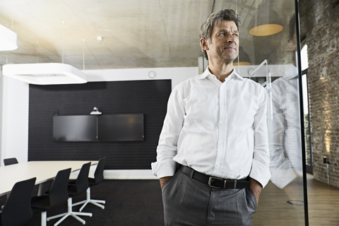 Porträt eines reifen Geschäftsmannes, der sich in einem modernen Konferenzraum gegen eine Glasscheibe lehnt, lizenzfreies Stockfoto