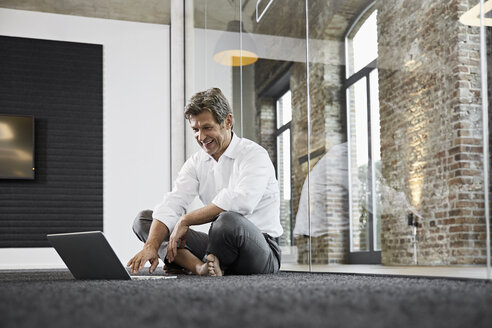 Barefoot businessman using laptop on floor in modern office - PDF01518