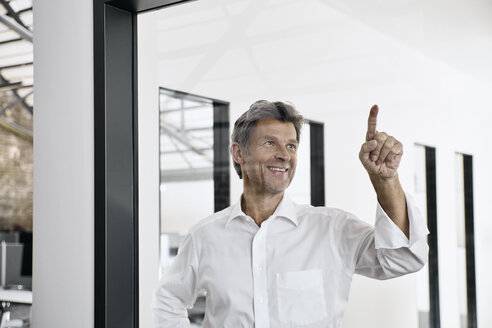 Smiling mature businessman touching glass pane in modern office - PDF01544