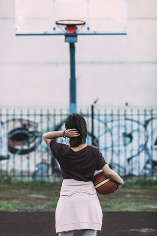 Rückansicht einer jungen Frau, die mit einem Basketball auf einem Platz im Freien steht, lizenzfreies Stockfoto