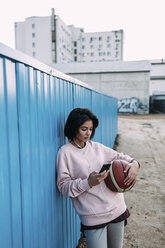 Junge Frau mit Basketball, Smartphone und Kopfhörern am Container - VPIF00338