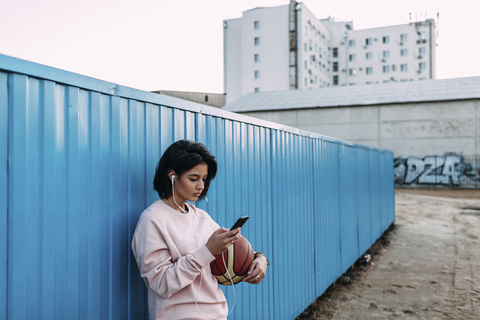 Junge Frau mit Basketball, Smartphone und Kopfhörern am Container, lizenzfreies Stockfoto