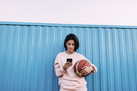 Junge Frau mit Basketball, Smartphone und Kopfhörern am Container, lizenzfreies Stockfoto