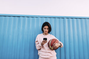 Junge Frau mit Basketball, Smartphone und Kopfhörern am Container - VPIF00341
