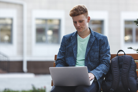 Rothaariger junger Mann sitzt auf einer Bank und benutzt einen Laptop, lizenzfreies Stockfoto