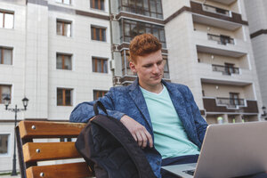 Rothaariger junger Mann sitzt auf einer Bank und benutzt einen Laptop - VPIF00350