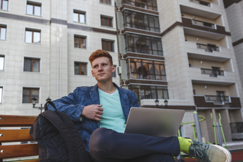 Rothaariger junger Mann sitzt mit Laptop auf einer Bank, lizenzfreies Stockfoto