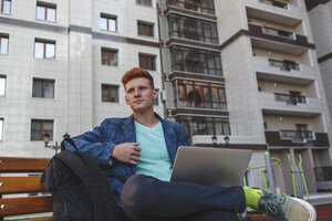 Rothaariger junger Mann sitzt mit Laptop auf einer Bank - VPIF00351