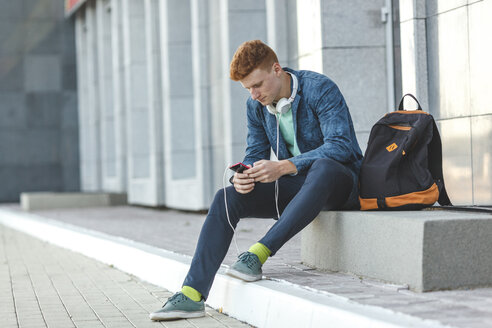 Rothaariger junger Mann sitzt mit Smartphone und Kopfhörern im Freien - VPIF00353