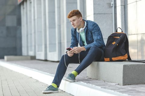 Rothaariger junger Mann sitzt mit Smartphone und Kopfhörern im Freien, lizenzfreies Stockfoto