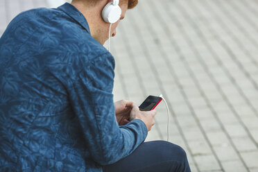 Junger Mann sitzt mit Smartphone und Kopfhörern im Freien - VPIF00355