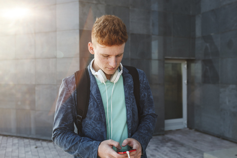 Rothaariger junger Mann im Freien mit Smartphone und Kopfhörern, lizenzfreies Stockfoto