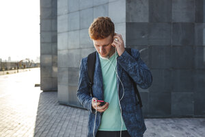 Rothaariger junger Mann im Freien mit Smartphone und Kopfhörern - VPIF00359