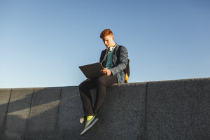 Rothaariger junger Mann sitzt an der Wand und benutzt einen Laptop - VPIF00363