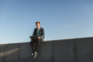 Lächelnder rothaariger junger Mann sitzt mit Laptop an der Wand - VPIF00364