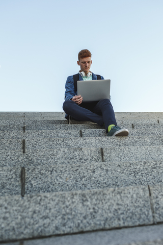 Rothaariger junger Mann sitzt auf einer Treppe und benutzt einen Laptop, lizenzfreies Stockfoto