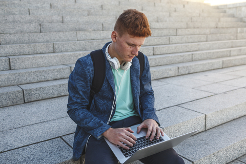 Redheaded young man sitting on stairs using laptop stock photo