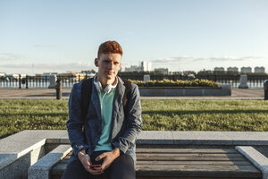 Porträt eines rothaarigen jungen Mannes, der mit Smartphone und Kopfhörern auf einer Bank sitzt - VPIF00369