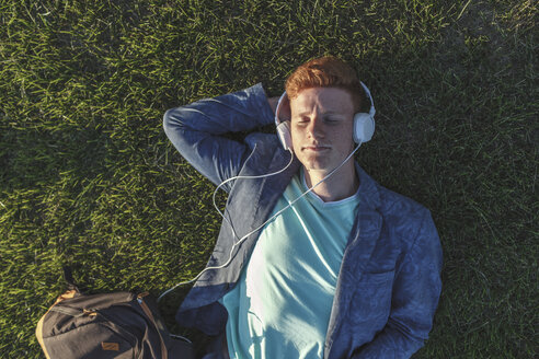 Rothaariger junger Mann mit Kopfhörern im Gras liegend - VPIF00372