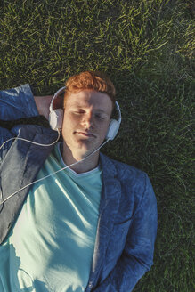 Rothaariger junger Mann mit Kopfhörern im Gras liegend - VPIF00373