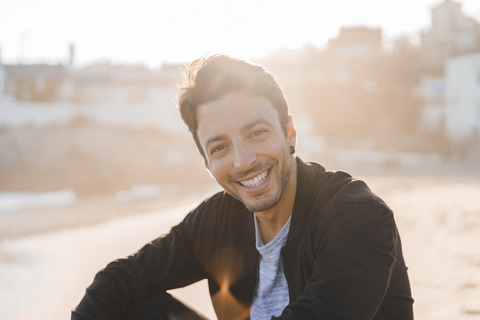 Portrait of smiling young man sitting on the beach at sunset stock photo