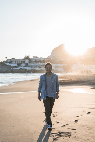 Glücklicher junger Mann, der bei Sonnenuntergang am Strand spazieren geht, lizenzfreies Stockfoto