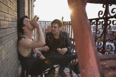 Zwei junge Männer sitzen auf einer Feuerleiter und trinken Bier, lizenzfreies Stockfoto