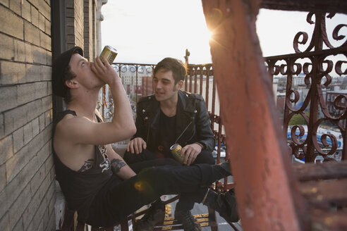 Zwei junge Männer sitzen auf einer Feuerleiter und trinken Bier - FSIF02656