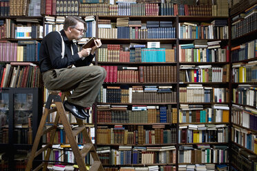 Ein Mann sitzt auf einer Leiter in einer Buchhandlung und liest ein Buch - FSIF02813