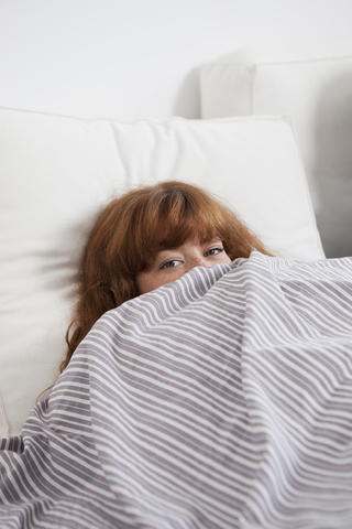 Eine Frau, die im Bett hinter einer Bettdecke hervorschaut, lizenzfreies Stockfoto