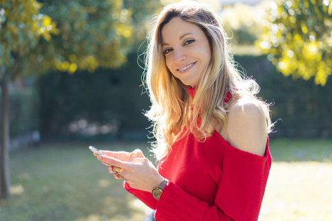 Porträt einer lächelnden jungen Frau, die in einem Garten ihr Smartphone benutzt, lizenzfreies Stockfoto