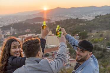 Spain, Barcelona, three happy friends with beer bottles on a hill overlooking the city at sunset - AFVF00222