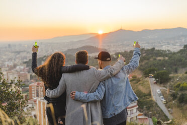 Spain, Barcelona, three friends with beer bottles embracing on a hill overlooking the city at sunset - AFVF00223