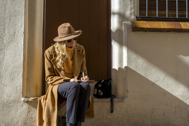 Modische junge Frau sitzt am Hauseingang und schreibt in ein Notizbuch - AFVF00239
