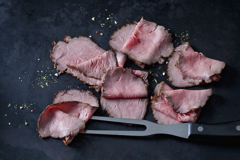 Sechs Scheiben Roastbeef, Kräuter und Fleischgabel auf dunklem Grund, lizenzfreies Stockfoto