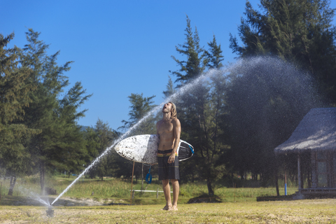 Young man with surfboard refreshing at sprinkler stock photo