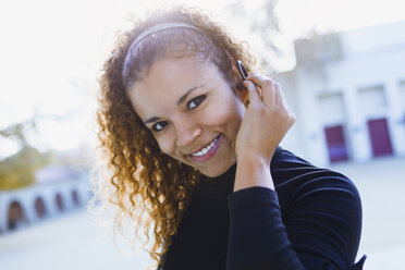 Portrait of smiling young woman outdoors putting on earphone - JSRF00001