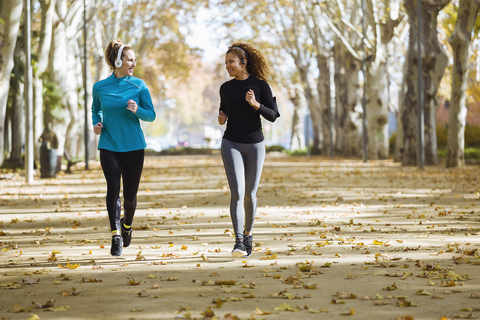 Two smiling young women running in park listening to music stock photo