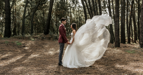 Bride with windswept wedding dress and groom standing in forest holding hands stock photo