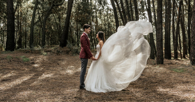 Braut mit windgepeitschtem Hochzeitskleid und Bräutigam stehen im Wald und halten sich an den Händen - DAPF00899