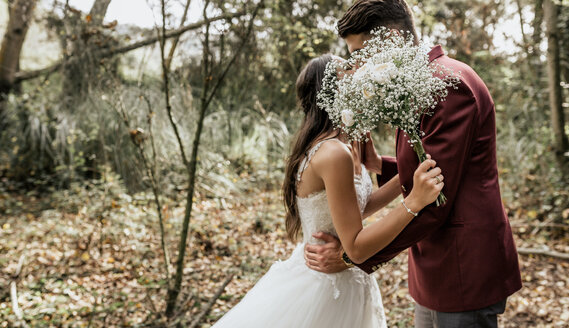 Unrecognizable bride and groom kissing in forest behind bouquet of flowers - DAPF00905