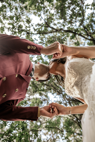 Braut und Bräutigam halten sich an den Händen und küssen sich unter einem Baum, lizenzfreies Stockfoto