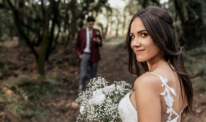 Porträt einer lächelnden Braut, die einen Blumenstrauß im Wald hält, mit dem Bräutigam im Hintergrund - DAPF00917